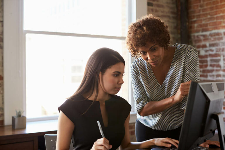 older female professional guiding young female professional using computer
