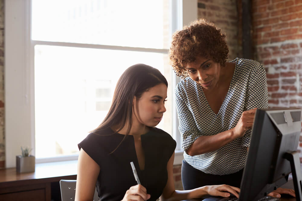 older female professional guiding young female professional using computer